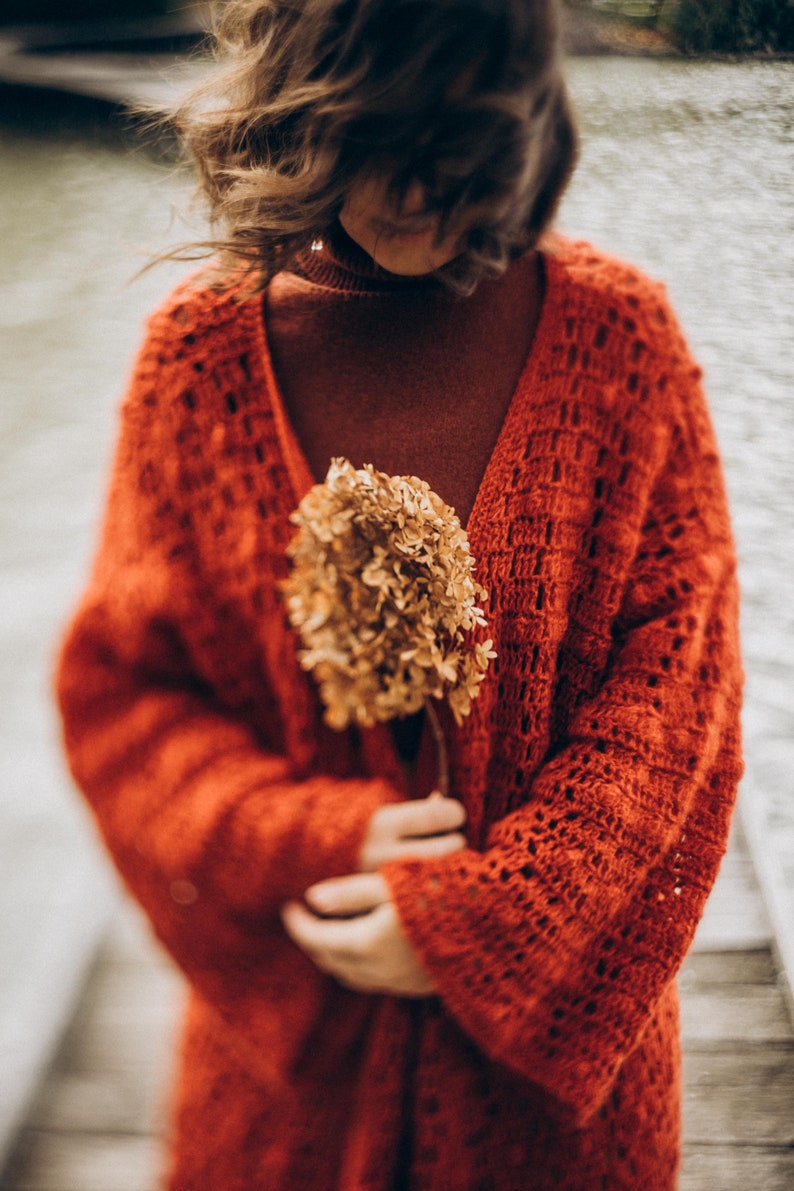 May include: A woman wearing a red crocheted cardigan holds a dried flower bouquet in front of her. The flower is a light brown colour and has a delicate texture.