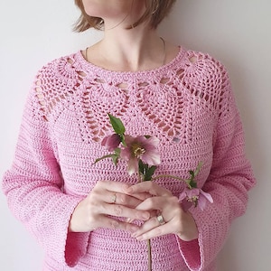 Op de afbeelding: Een roze gebreide trui met een kantkraag en lange mouwen. De trui wordt vastgehouden door een persoon, en er zijn roze bloemen op de voorgrond.