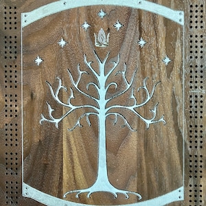 Könnte beinhalten: Ein Holzbrett mit einer silbernen Metallplatte, auf der ein weißer Baum mit einer Krone obenauf abgebildet ist. Der Baum ist von sieben Sternen umgeben. Das Brett hat ein Gitter aus Löchern.