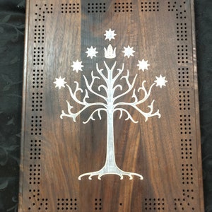 Könnte beinhalten: Ein Holzspielbrett mit einem weißen Baum- und Sternenmotiv. Das Brett hat eine dunkelbraune Holzoptik und einen weißen Rand. Der Baum ist eine stilisierte Version des Weißen Baumes von Gondor aus Der Herr der Ringe.