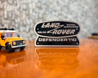 May include: A black and silver metal plaque featuring the text LAND ROVER, SOLIHULL WARWICKSHIRE, ENGLAND, and DEFENDER 110, positioned next to a small yellow toy vehicle constructed from plastic building blocks with a black roof rack.