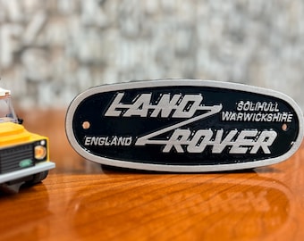Puede incluir: Un emblema ovalado de metal de Land Rover con fondo negro y letras plateadas que dicen Land Rover, Solihull Warwickshire y England. Al lado hay un pequeño vehículo de juguete construido con bloques de construcción de plástico.