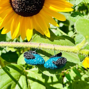 Puede incluir: Un par de pendientes de ganchillo en azul y negro. Los pendientes cuelgan de un tallo de girasol.