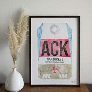 May include: A framed watercolor print of a luggage tag with the airport code "ACK" for Nantucket Memorial Airport. The tag is painted in shades of blue, red, and gray with a small red airplane icon.