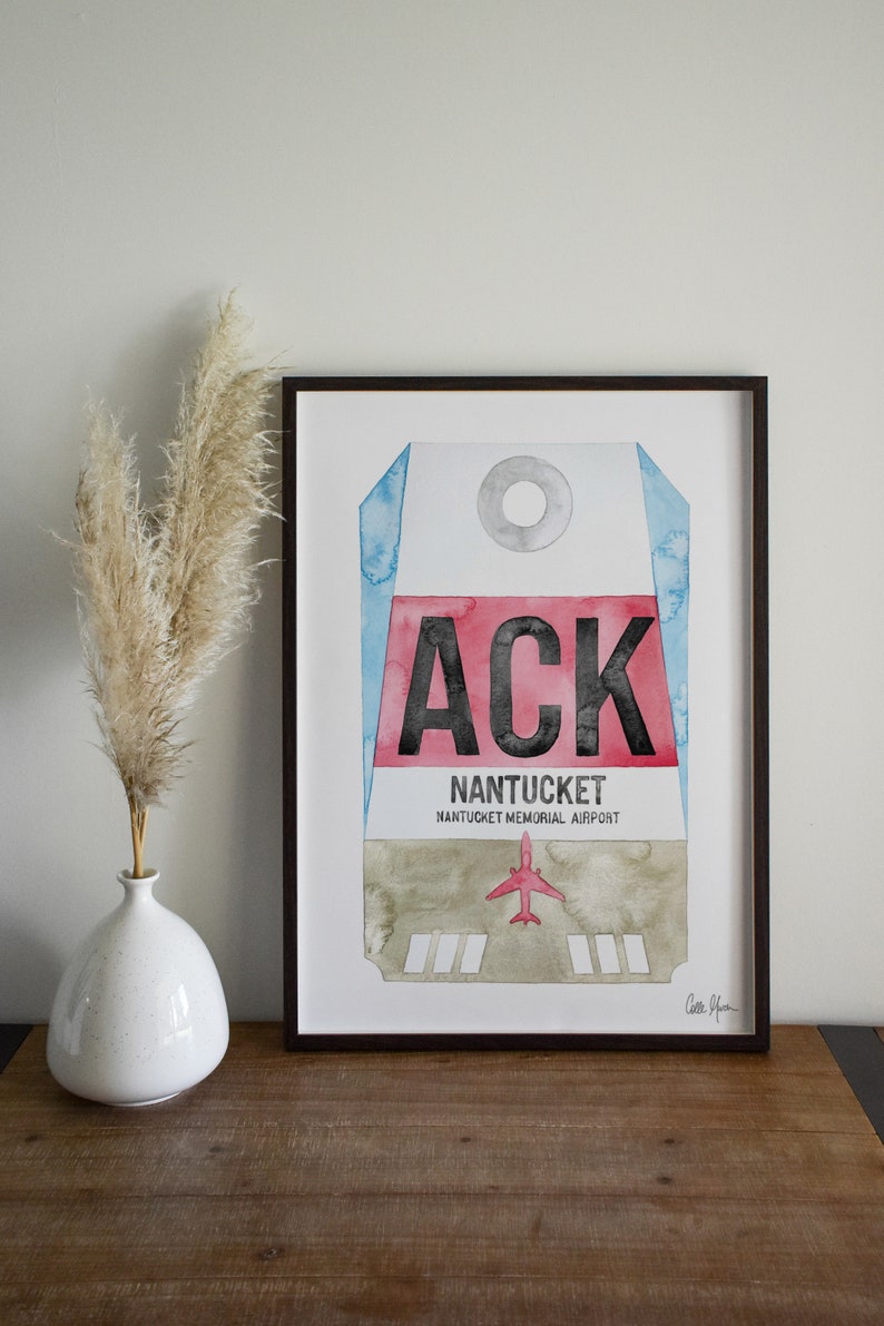 May include: A framed watercolor print of a luggage tag with the airport code "ACK" for Nantucket Memorial Airport. The tag is painted in shades of blue, red, and gray with a small red airplane icon.