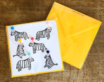 May include: A greeting card featuring six cartoon zebras with party hats and colorful pom-poms. The card is in a clear plastic sleeve, next to a bright yellow envelope. The envelope has the text "COMPOSTABLE BAG".
