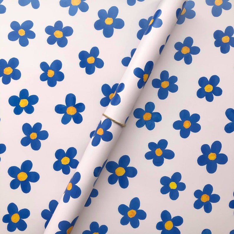 May include: White wrapping paper with a repeating pattern of blue flowers with yellow centers. A roll of the wrapping paper is partially unrolled and lying on top of the paper.