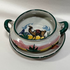 May include: A white ceramic bowl with green handles and a painted design of a camel with a rider. The bowl has a matching saucer with a similar design.