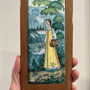 Puede incluir: Azulejo de cerámica enmarcado que representa a una mujer con un vestido amarillo que lleva una cesta en una escena de bosque. La obra de arte tiene un estilo de arte popular y un marco de madera. La paleta de colores incluye verdes, azules y amarillos.
