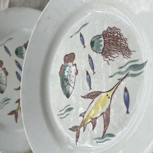 Puede incluir: Dos platos de cerámica blanca con diseños de vida marina pintados a mano. Uno presenta un pez espada amarillo, una medusa y pequeños peces azules. El otro plato tiene un diseño similar con un pez diferente. Los platos tienen una estética rústica y vintage.