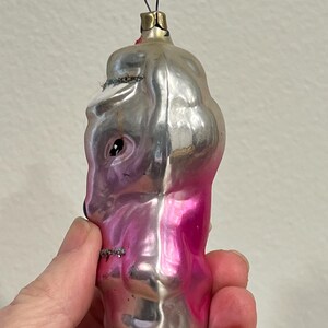 Puede incluir: Un adorno navide&ntilde;o de vidrio vintage con forma de persona con acabado rosa y plateado. El adorno tiene una cara plateada con un ojo negro y un cuerpo rosa.