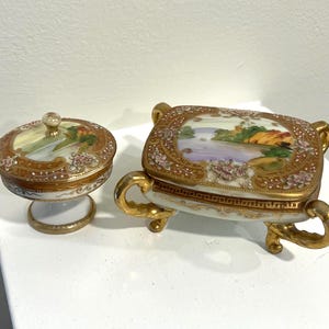 Puede incluir: Dos joyeros antiguos de porcelana. Uno es un pequeño tarro redondo con tapa sobre una base de pedestal. El otro es una caja rectangular con tapa, sostenida por cuatro patas curvas. Ambos presentan escenas de paisajes pintadas a mano y detalles dorados.