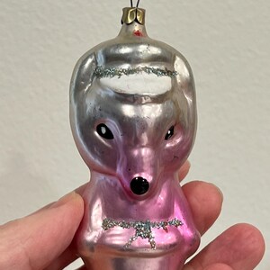 Puede incluir: Un adorno de Navidad de vidrio rosa con forma de oso con un sombrero blanco y una nariz negra. El adorno tiene detalles de purpurina.