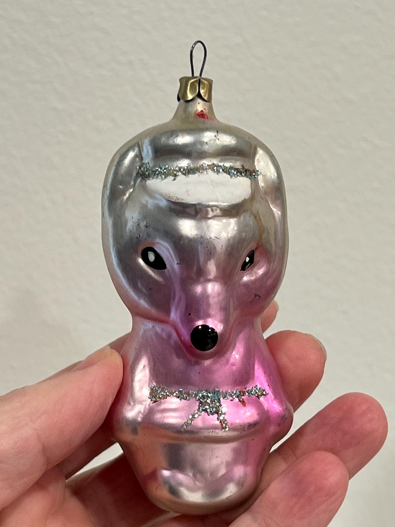 Puede incluir: Un adorno de Navidad de vidrio rosa con forma de oso con un sombrero blanco y una nariz negra. El adorno tiene detalles de purpurina.