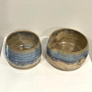 May include: Two handmade ceramic bowls. The bowls are a mix of tan and blue, with a layered, textured appearance. The bowl on the right has a spout. The bowls are on a white surface.