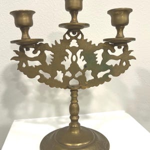 Puede incluir: Un candelabro de latón con tres candeleros. La pieza decorativa presenta un diseño calado ornamentado con un tallo central y una base redonda. El candelabro es de color dorado cálido.