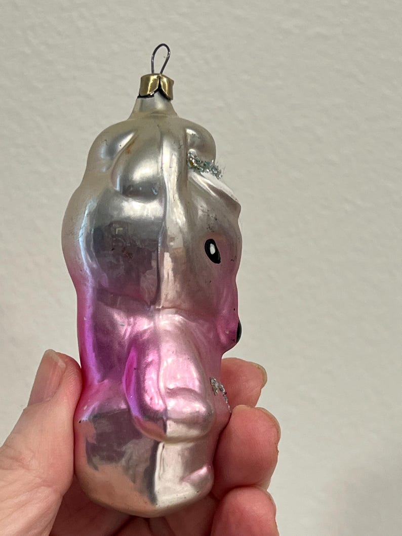 Puede incluir: Un adorno de Navidad de vidrio rosa y plateado con forma de oso con sombrero. El adorno tiene un lazo de alambre plateado para colgar.