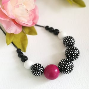 Peut inclure: Un collier de perles en bois avec des pois noirs et blancs et une grosse perle rose fuchsia. Le collier est composé de perles en bois et est enfilé sur une chaîne noire.