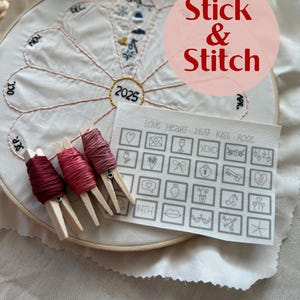 May include: A close-up of a white embroidery hoop with a floral design and the year 2025 embroidered in the center. The hoop is surrounded by a pink and white fabric with the words "Stick & Stitch" printed in red. There are four spools of red embroidery floss with wooden clothespins attached to them. A sheet of paper with a grid of small squares containing various embroidery designs is visible in the lower right corner.