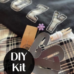May include: DIY kit for creating a personalized "Cozy" patch with black and white plaid fabric, gray felt letters, and a small purple flower.