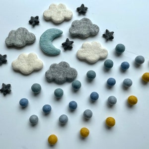 May include: A selection of felted wool shapes, including grey and white clouds, dark grey stars, a light blue crescent moon, and blue and yellow spheres. These elements are arranged on a white surface, suitable for a nursery or craft project.