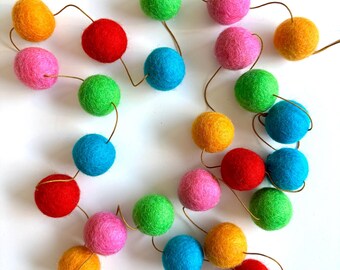 Puede incluir: Una colorida guirnalda de bolas de fieltro en tonos naranja, rosa, rojo, verde y azul. Las bolas redondas están unidas por un fino alambre dorado. La guirnalda está dispuesta en un patrón circular sobre un fondo blanco.