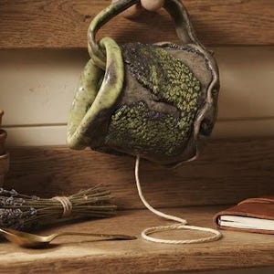 May include: A handmade ceramic yarn bowl with a mossy green and dark brown textured exterior, featuring a side cutout for yarn. The bowl hangs from a wooden peg, with a single strand of light-colored yarn extending downward from the opening.