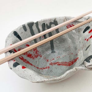 May include: A handmade ceramic bowl with a speckled gray finish, accented with red and black paint. Two wooden chopsticks rest across the bowl, suggesting a culinary theme.