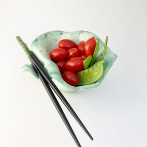 May include: A small, green and white ceramic bowl with a ruffled edge, filled with red cherry tomatoes and green pea pods. Two black chopsticks are placed next to the bowl.