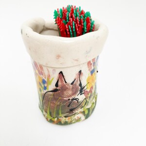 May include: A handmade ceramic container with a painted fox design, filled with red and green toothpicks. The container has a cream-colored rim and is decorated with colorful flowers.