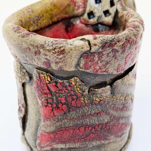 May include: A handmade ceramic cup with a textured surface. The cup features a combination of red, yellow, and brown colors. The interior is a deep red, and the exterior has a cracked, textured design. The cup has a unique handle with square cutouts.