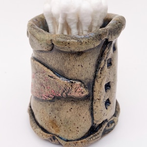 May include: A small, handmade ceramic jar with a textured, brown and green glaze. The jar has a pink and brown design on the side and is filled with cotton swabs.