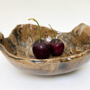 May include: A handcrafted, organic-shaped ceramic bowl in shades of brown and tan, with a textured surface and decorative details. Two ripe, dark red cherries with green stems rest inside the bowl. The bowl is on a white background.
