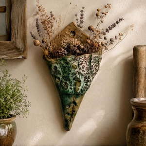 May include: A triangular ceramic wall pocket planter featuring a mottled green and brown glaze with a textured, speckled surface. The vessel is mounted to the wall and contains an arrangement of dried lavender and various brown botanical stems.