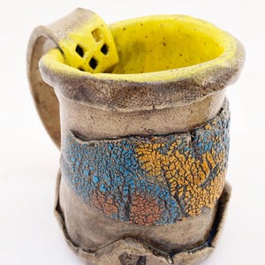 May include: A handmade ceramic mug with a textured, earthy brown exterior and a bright yellow interior. The side features a decorative band with a cracked, multi-colored blue and orange glaze. A wide, flat handle includes a small square cutout.