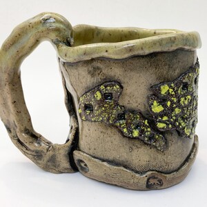 May include: Handmade ceramic mug with a unique, textured design. The mug features a tree branch-like handle and a textured, earthy-toned body with accents of green and black. The rim and handle have a light green glaze.