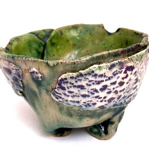 May include: A hand-built ceramic bowl with an irregular, organic shape and a textured surface. The bowl features a mottled green glaze with prominent patches of white and dark purple speckled patterns on the exterior and a deep green interior.