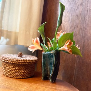 May include: A small, dark blue ceramic vase with a textured surface holds a bouquet of light orange flowers and green leaves. A small, round, woven basket with a lid sits on a wooden table.