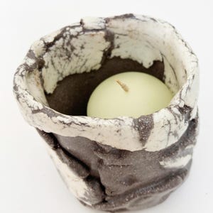 May include: A handmade ceramic candle holder with a textured, dark brown exterior and a white and gray marbled rim. Inside, a round, pale green candle sits nestled within. The candle holder is designed to hold a single tea light.