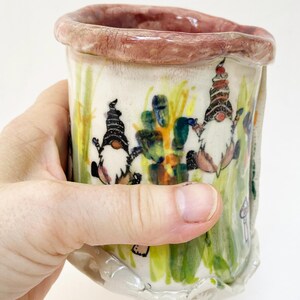 May include: Hand-painted ceramic mug featuring a whimsical design of gnomes in a garden setting. The mug has a cream-colored base with colorful illustrations of gnomes, plants, and mushrooms. The rim is a reddish-brown color.
