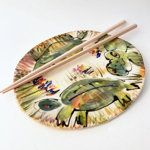 May include: A hand-painted ceramic plate with a turtle design in green, orange, and blue. Two wooden chopsticks rest on the plate. The plate has a cream-colored background with floral accents.