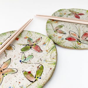 May include: Two ceramic plates with dragonfly designs and a pair of wooden chopsticks. The plates feature a speckled white background with hand-painted dragonflies in red, green, and pink. Green and yellow grass and small flowers are also painted on the plates.