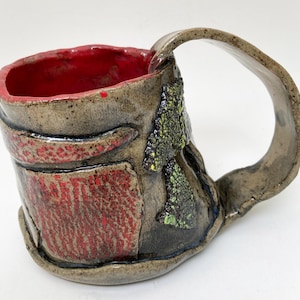 May include: Handmade ceramic mug with a textured, earthy design. The interior is a vibrant red, contrasting with the exterior's muted tones. Features a large, curved handle and decorative green and yellow accents. A unique piece for enjoying beverages.