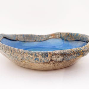 May include: A shallow ceramic bowl with an irregular, organic rim. The interior features a smooth, vibrant blue glaze, while the exterior and the thick, textured rim display a mottled, earthy brown and blue finish with a rough, granular surface.