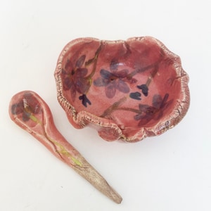 May include: A pink ceramic bowl with a floral design and a matching spoon. The bowl has a scalloped edge and the spoon has a curved handle.