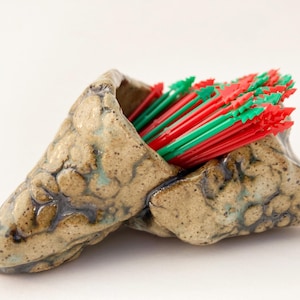 May include: A ceramic holder with a rough, textured surface in shades of brown and green, holding a bunch of red and green plastic cocktail sticks.