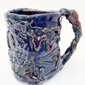 May include: A handmade ceramic mug with a glossy deep blue glaze and textured, raised surface patterns. The mug features a thick, irregular handle with red speckled accents that match the textured details on the body of the vessel.