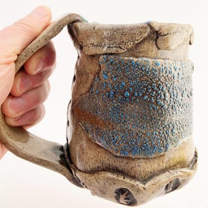 May include: A ceramic mug with a textured, earthy brown surface and a prominent, rough-hewn handle. The body features a central panel with a raised, bubbly blue glaze that contrasts with the smooth, mottled tan clay finish of the rest of the vessel.