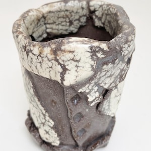 May include: A handmade ceramic cup with a unique, textured design. The cup features a dark brown base with a cream-colored, crackled glaze around the rim and accents. The cup has a slightly irregular shape, adding to its handcrafted appeal.