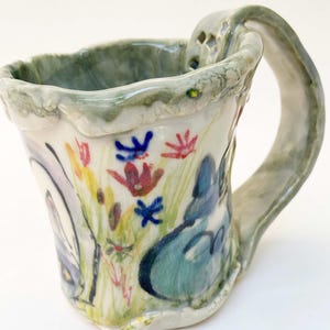 May include: Hand-painted ceramic mug with a whimsical design. The mug features a white base with colorful floral and abstract illustrations in blue, red, yellow, and green. The handle and rim have a textured, light green finish. A unique, artistic piece.